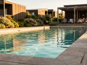 Glinsterende buitenzwembad bij een modern vakantiepark, omgeven door houten cabins, weelderig groen en een overdekt terras met ligstoelen.