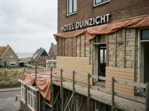 Verouderde hotelfaçade in renovatie met steigers, contrast tussen moderne panelen en origineel metselwerk, kuststad op de achtergrond.
