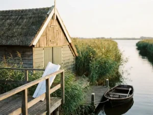Houten recreatiewoning met vlonder aan rustig waterkanaal, omgeven door rietkragen, met opgerolde bouwtekening op de reling in middagzon.